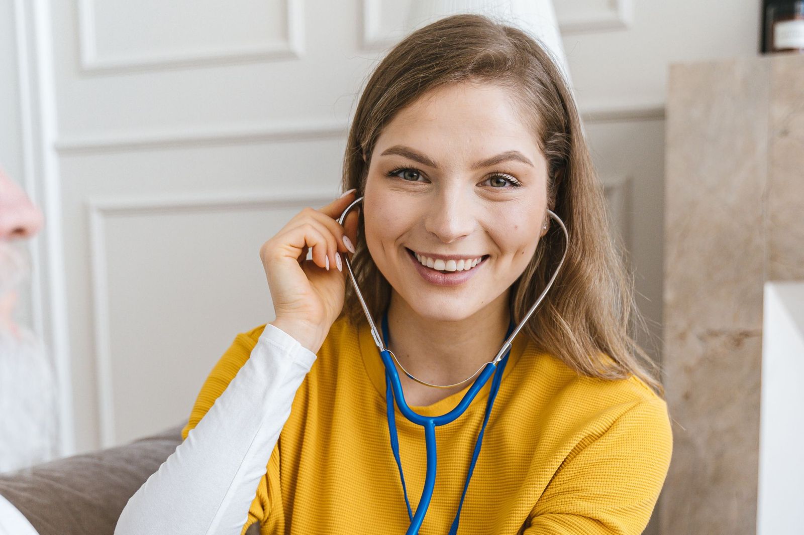 A smiling nurse in a yellow top listens with a stethoscope during a home visit.