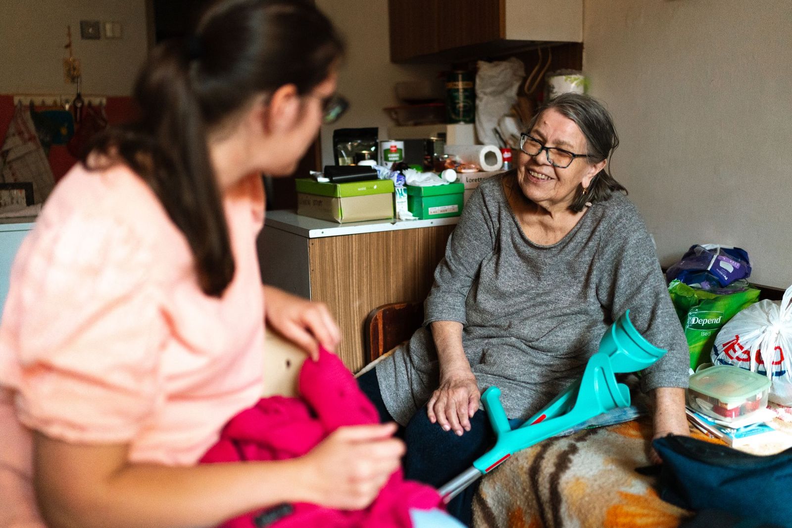 A caregiver helps an older woman sort through clothing in her cozy home as they chat.