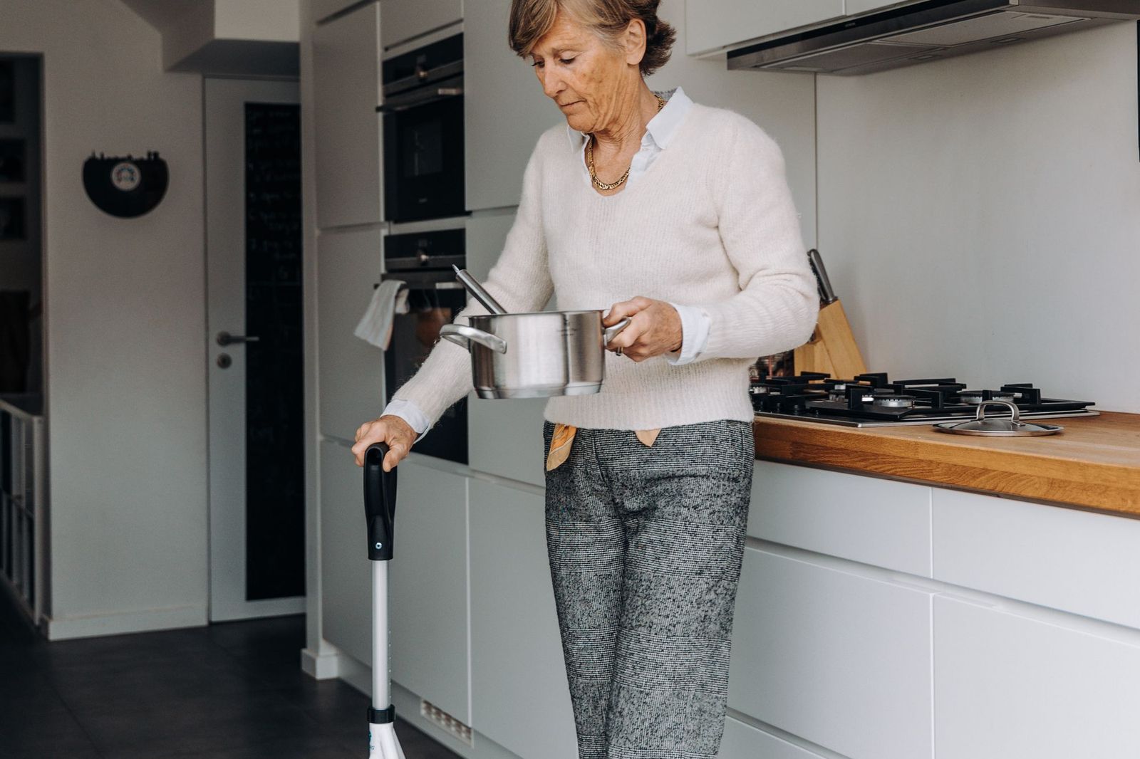 An older woman uses a rolling walker to move around her kitchen while carrying a cooking pot.