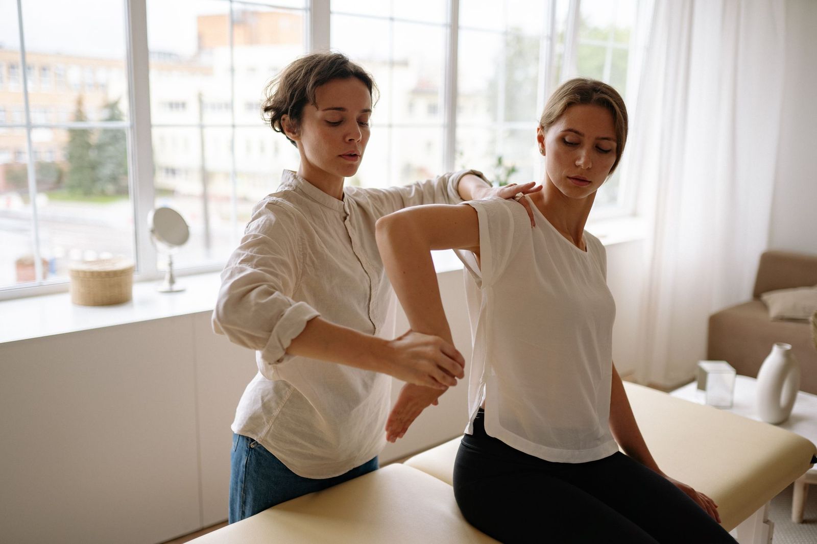 A physical therapist gently guides a woman’s arm through a shoulder exercise in a bright room.