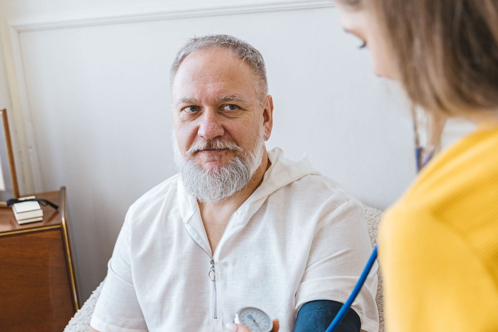A nurse in a yellow top takes an older man’s blood pressure while he sits in a comfortable armchair at home.